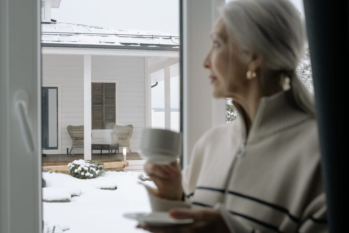 An older woman holding a cup and looking out of a window at a snowy porch