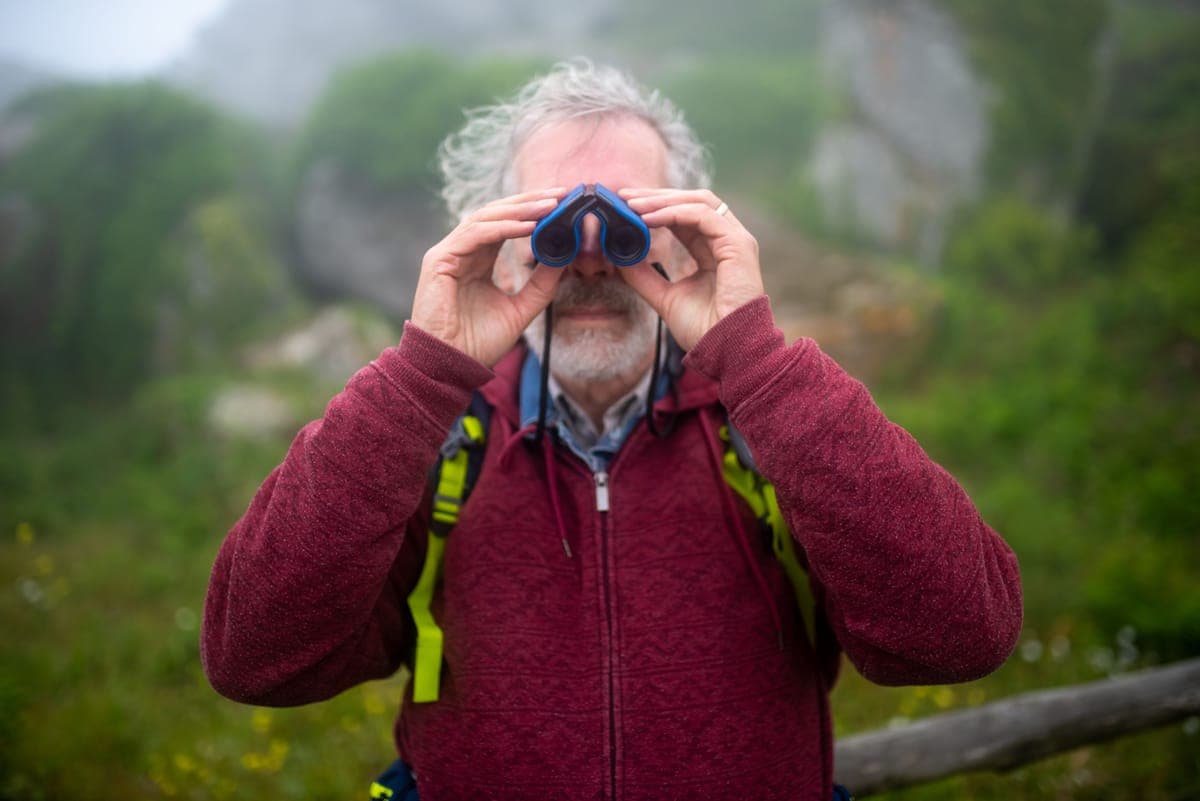 An older man walking in green countryside with binoculars